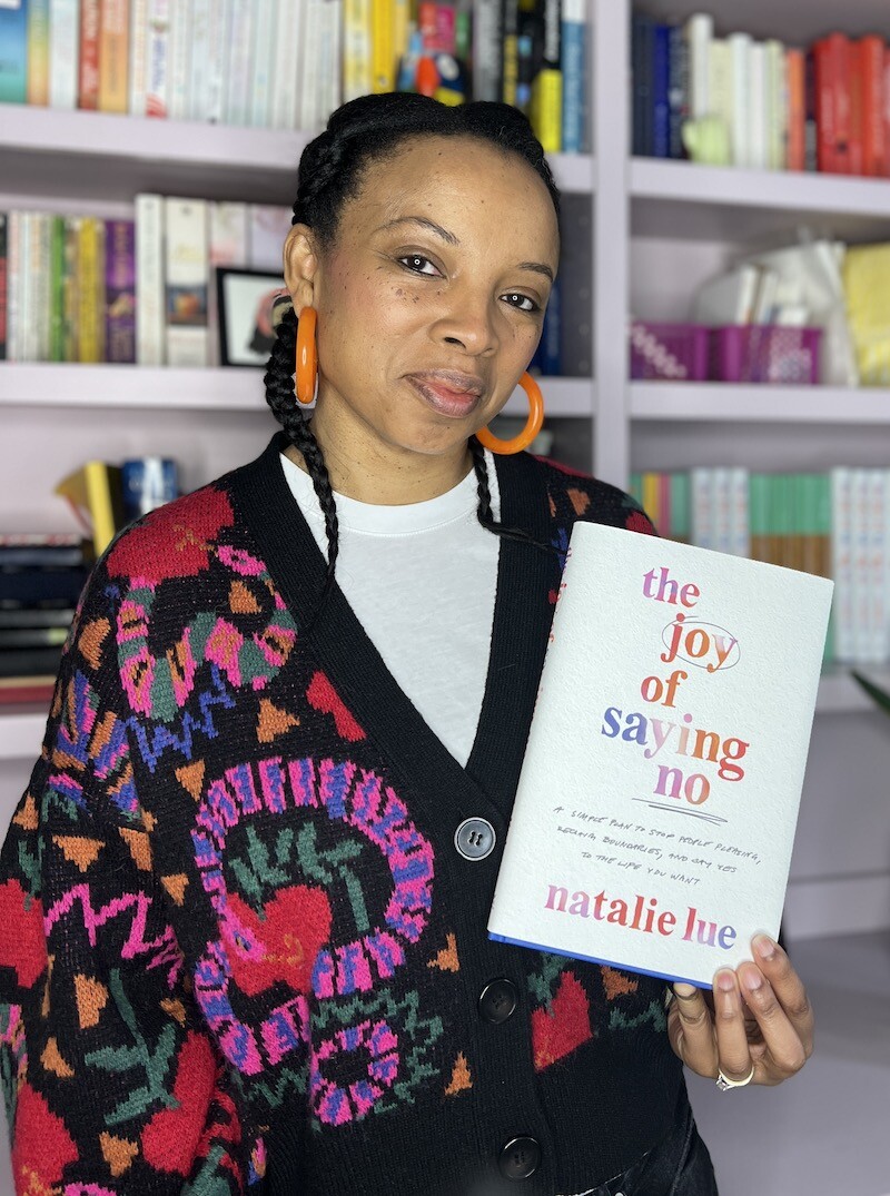 Natalie Lue standing in her studio space with her hair in two braids and wearing large orange hoops holding a copy of her book The Joy of Saying No