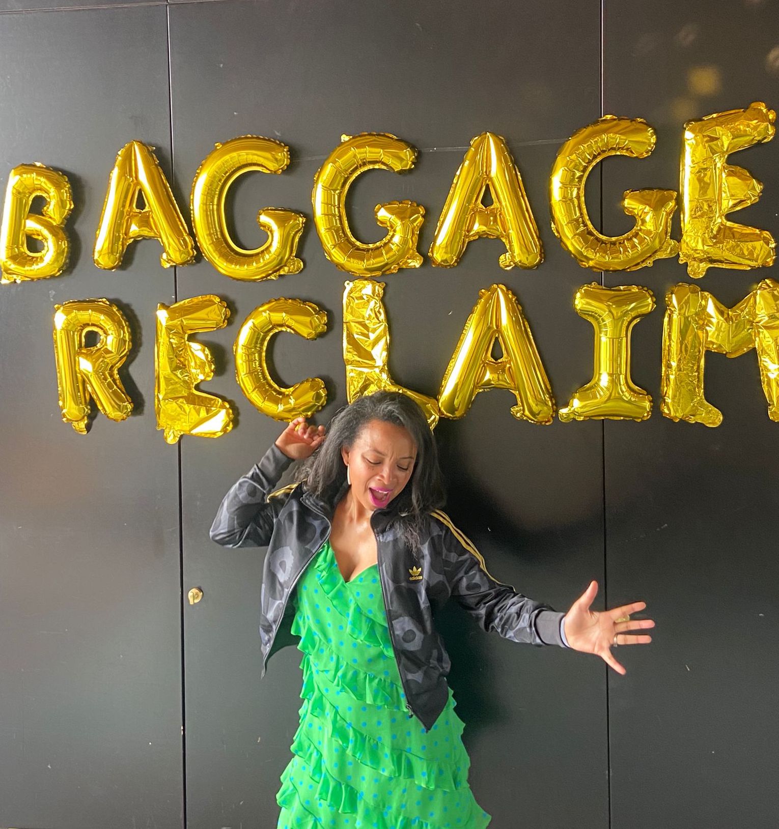 Natalie Lue in green polka dot dress and Marimekko Adidas jacket under gold, 18th birthday balloons spelling Baggage Reclaim