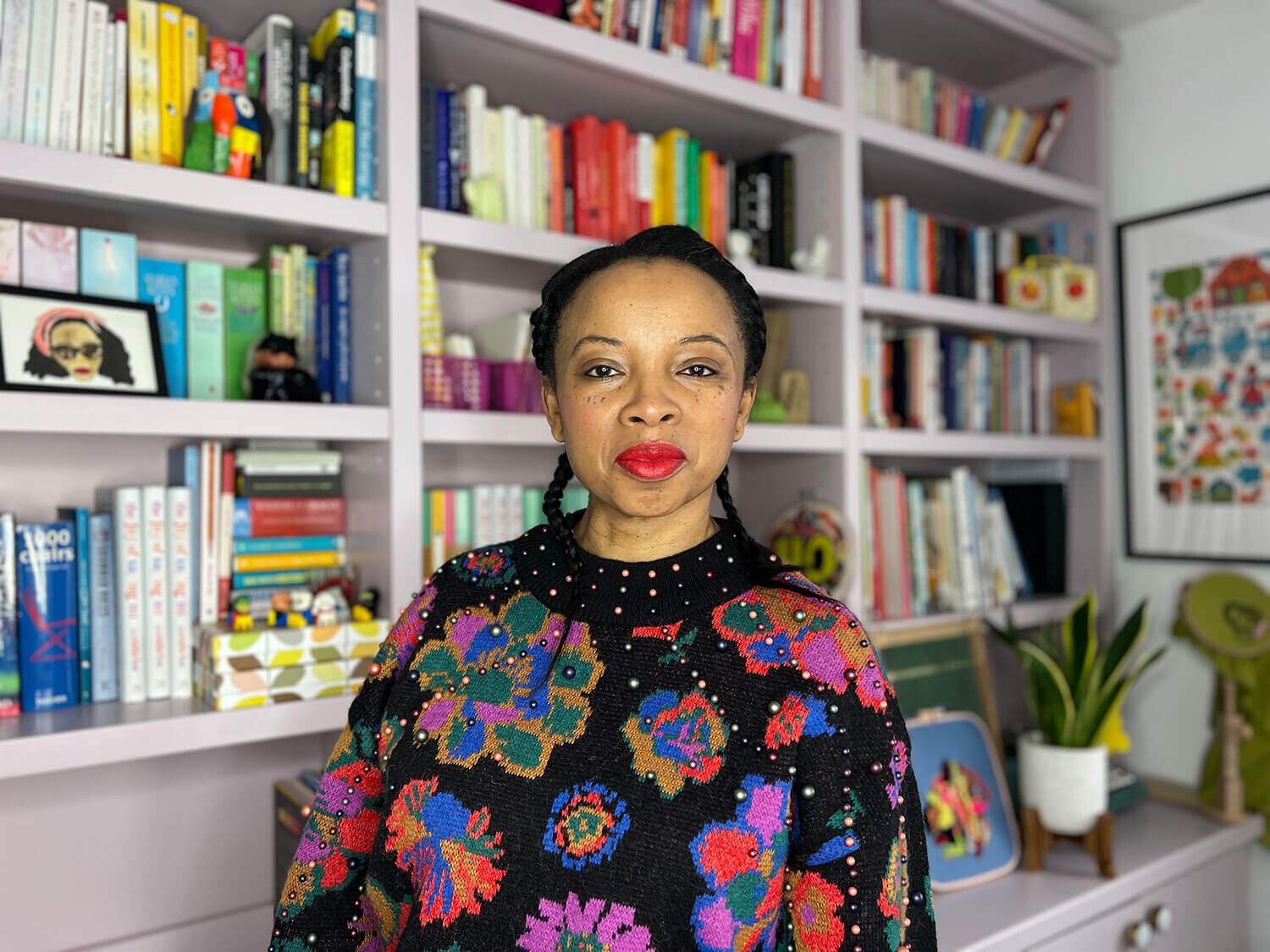 Natalie Lue standing in her studio with her books in the background wearing a Farm Rio jumper, red lipstick, and with her hair in two braids